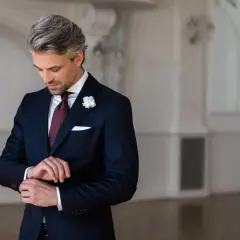 Mężczyzna w granatowym garniturze, białej koszuli i bordowym krawacie z białym kwiatem w butonierce.